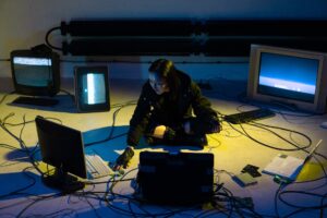 a woman in black jacket sitting on the floor while surrounded with televisions and computer