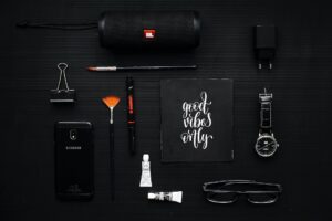 close up photography of smartphone beside binder clip speaker and eyeglasses on black textile